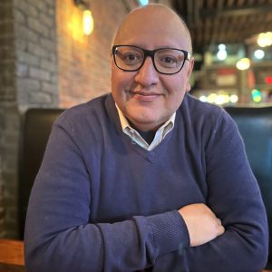Portrait of Mauricio Mendoza, a bald man wearing glasses and a navy sweater over a collared shirt, seated indoors and smiling warmly in a softly lit setting.