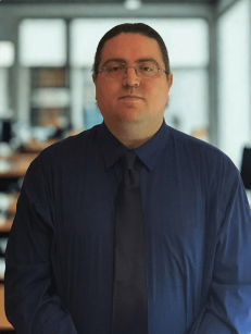 Portrait of Michael Salen standing indoors against a softly blurred office background. He is facing the camera with a neutral expression, wearing glasses, a dark blue dress shirt, and a matching navy tie, with his dark hair pulled back