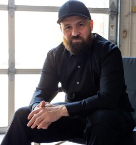 Kris Rivenburgh wearing dark pants and dress shirt along with a black baseball hat looking ahead in a professional style photo.