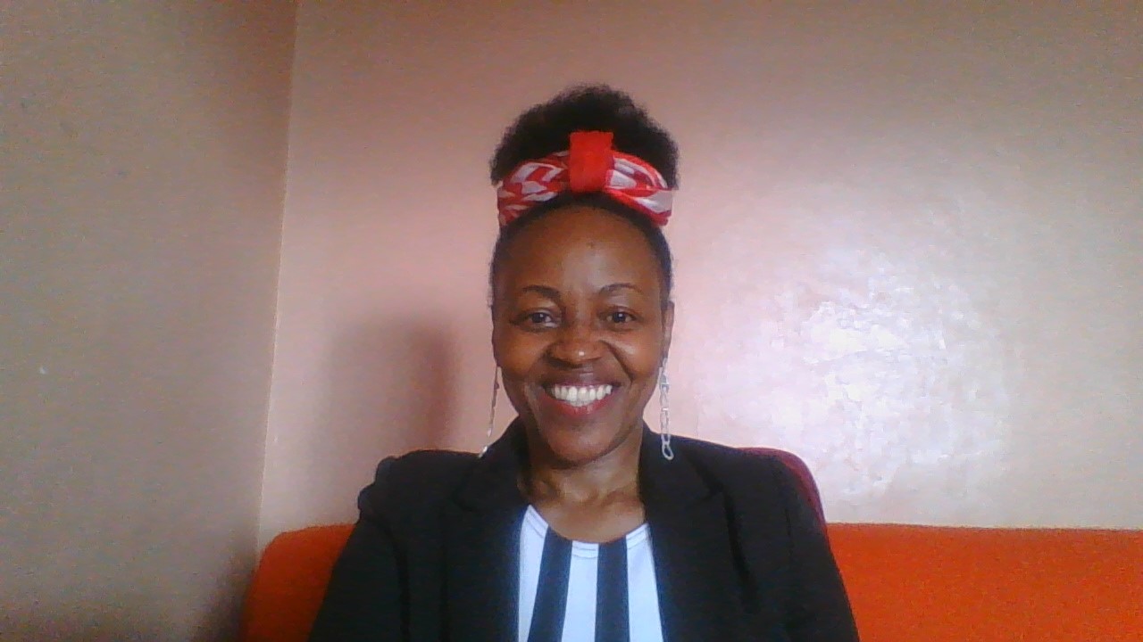 Professional headshot of a smiling Ruth Kaggwa wearing a black blazer and a red patterned headband.
